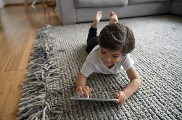 Child playing on a gray patterned area rug with a tablet in a bright living room